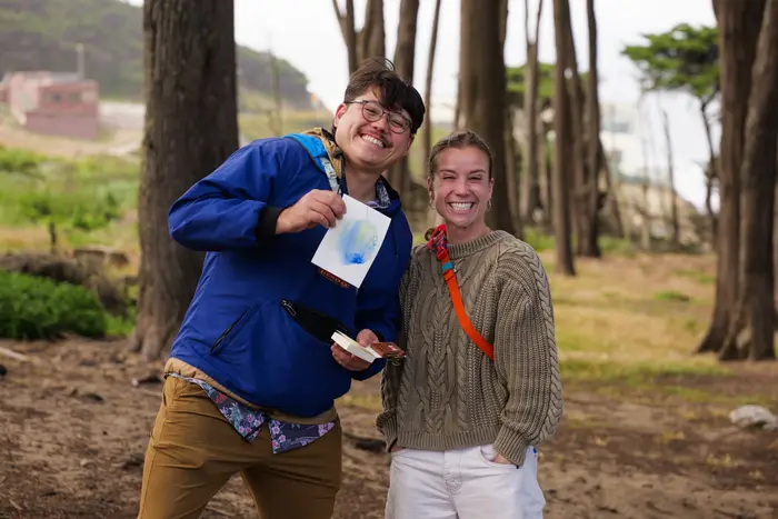 Two smiling watercolor workshop participants holding up their drawings among the trees.