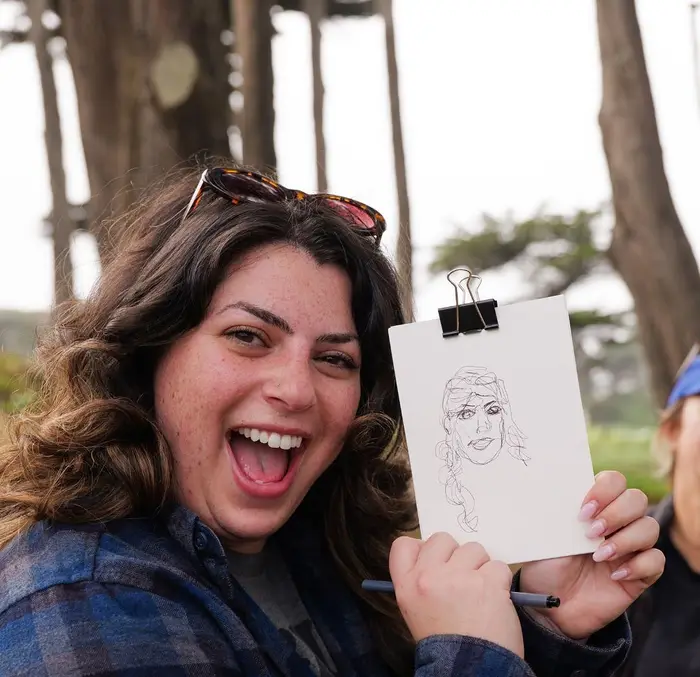 A workshop participant laughing as she holds up the blind contour portrait she drew in pen, cypress trees behind her.