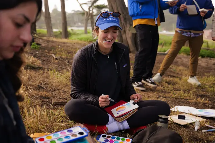 Participants seated together among coastal trees, working with watercolor palettes and sketchbooks.