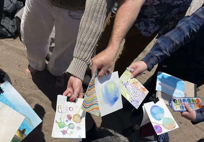 Looking down at a circle of hands on the beach, each holding out a finished watercolor page to share with the group at the end of the walk.