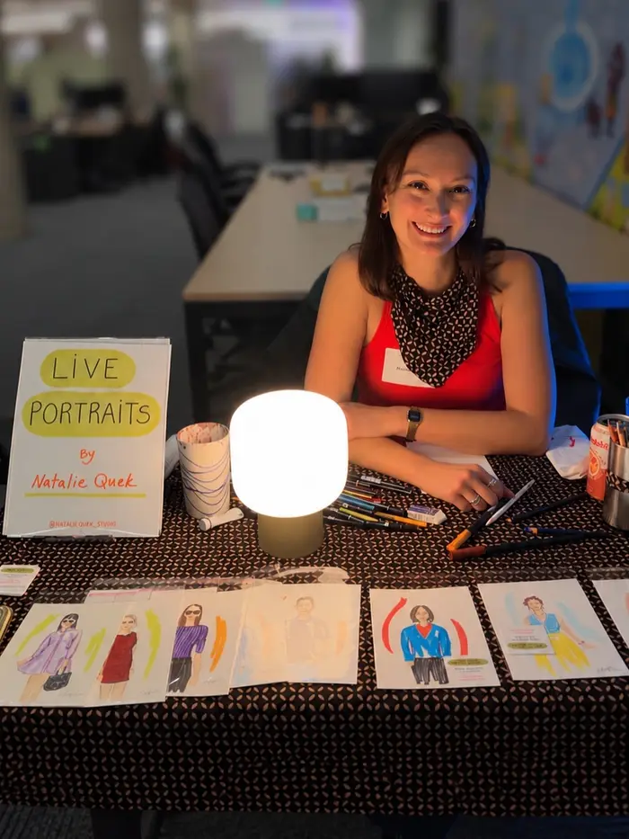 Natalie smiling at her live portrait table, with colorful guest illustrations and art supplies on display