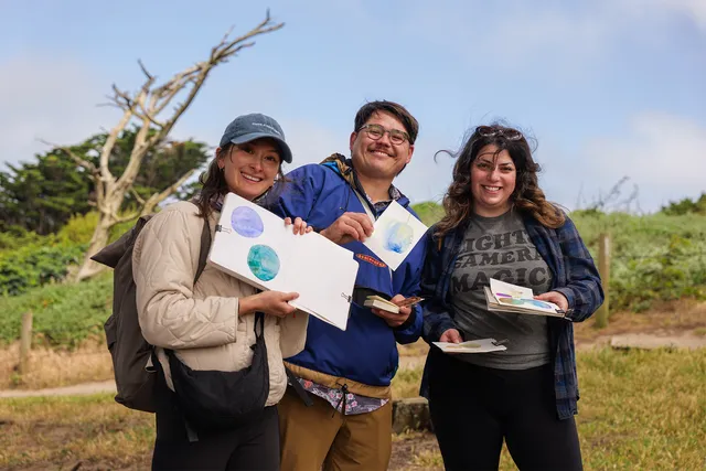 Three watercolor hike participants smiling and holding up their paintings outdoors