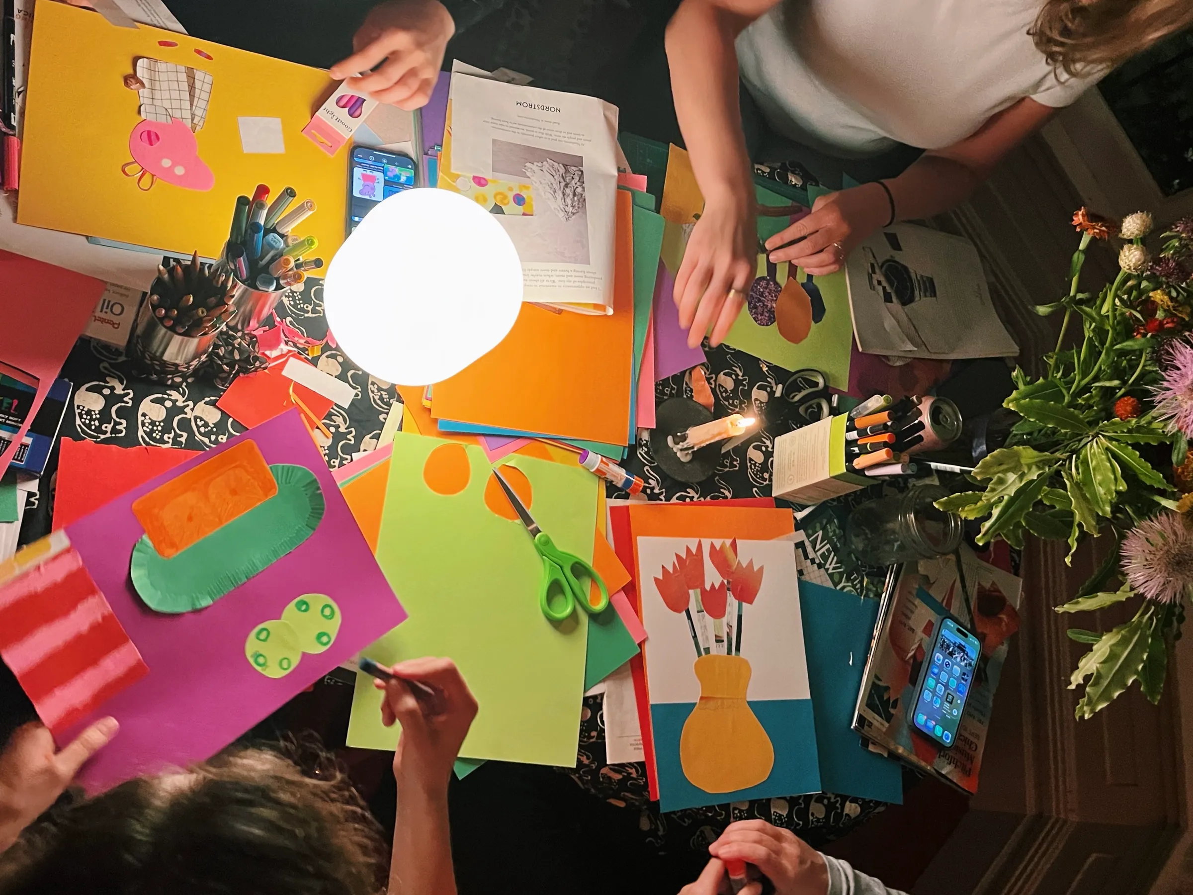 Overhead view of a collage workshop table with colorful paper artwork, scissors, drawing materials, and flowers